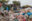 A village street is completely filled with plastic rubbish and bags of all colours and sizes, which is piled high on the right side of the road. A small girl is walking over rubbish towards the shack-link buildings just beyond.